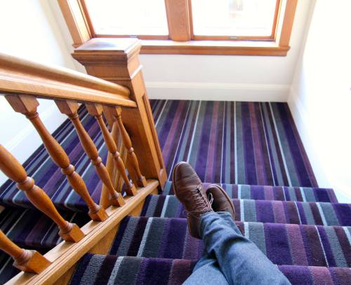 A person's foot in a brown shoe rests on striped carpeted stairs, with a wooden railing and a window in the background.