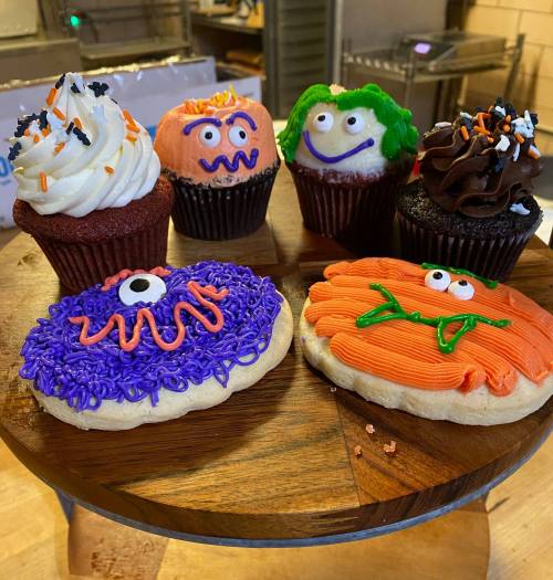 A wooden platter displays colorful Halloween-themed cupcakes and cookies, featuring fun monster designs and vibrant frosting.