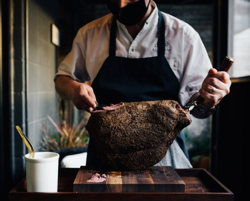 A chef in a mask carves a large piece of smoked meat on a wooden board, with a small cup nearby.