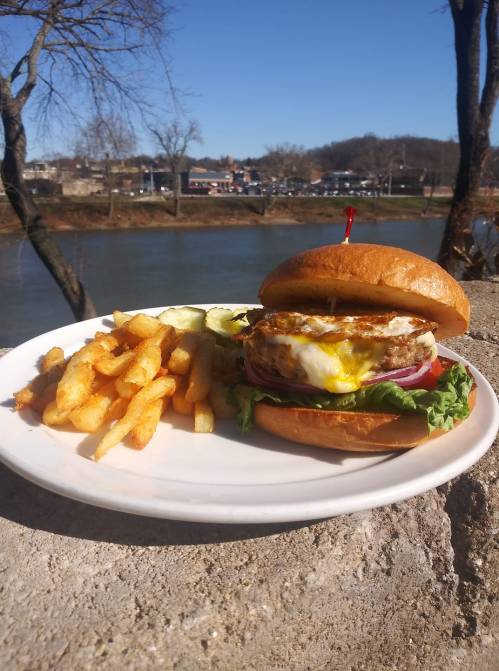 A juicy burger with a fried egg, lettuce, and tomato, served with crispy fries, against a scenic riverside backdrop.