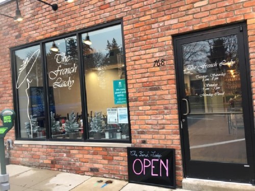 Exterior of "The French Lady" shop with large windows, brick wall, and an "OPEN" sign on the sidewalk.