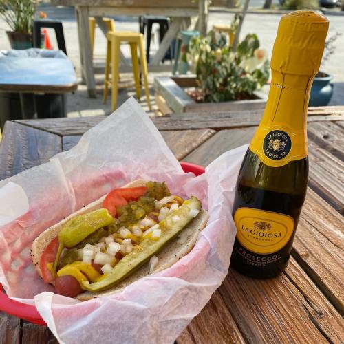 A hot dog topped with mustard, relish, onions, and peppers, next to a bottle of Prosecco on a wooden table.