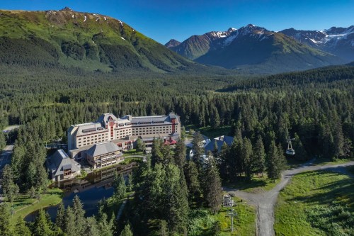 Aerial view of a large hotel surrounded by lush forests and mountains under a clear blue sky.
