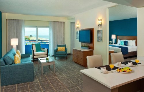 A modern hotel suite featuring a living area, kitchenette, and a bedroom with ocean views. Bright and inviting decor.