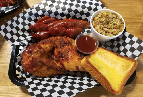 A plate of grilled chicken, sausage, rice, and toasted bread served with a small cup of sauce on a checkered tray.