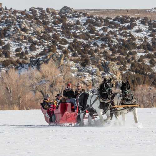 A horse-drawn sleigh carries passengers across a snowy landscape with rocky hills in the background.