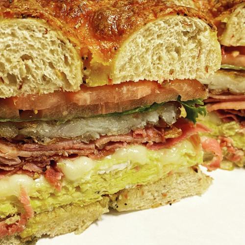 Close-up of a layered sandwich with meats, cheese, lettuce, tomatoes, and pickles on a toasted bread roll.