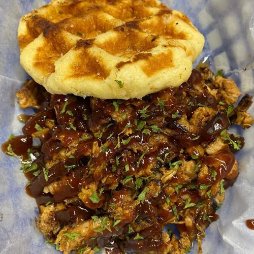 A plate featuring a golden waffle topped with shredded chicken and drizzled with barbecue sauce, garnished with herbs.