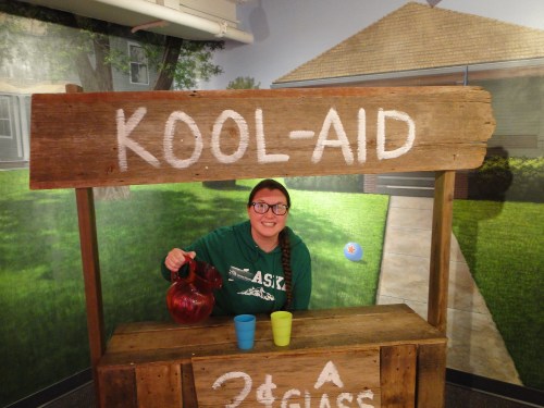 A person smiles behind a wooden Kool-Aid stand, holding a pitcher with colorful cups in front.