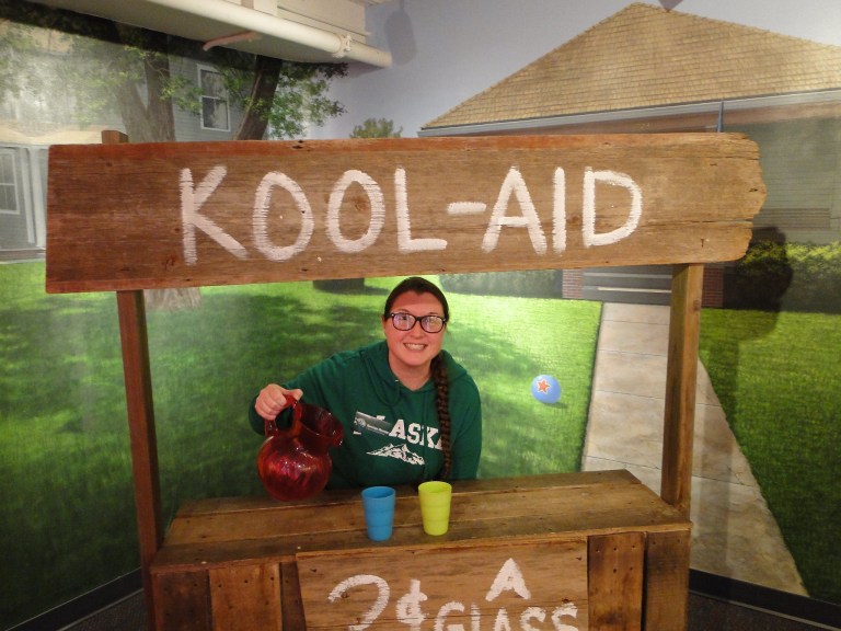 A person smiles behind a wooden Kool-Aid stand, holding a pitcher with colorful cups in front.