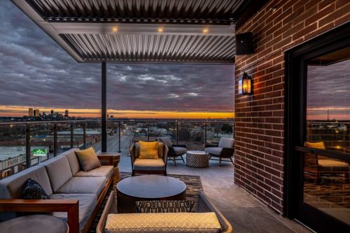 A modern balcony with seating, overlooking a city skyline at sunset with dramatic clouds.