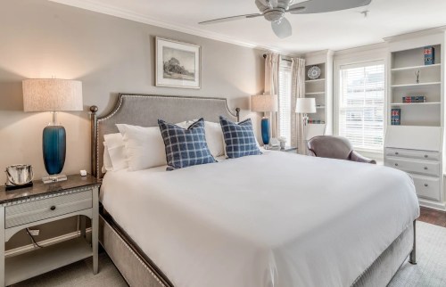 Cozy bedroom featuring a large bed, decorative pillows, bedside lamps, and a bookshelf with a chair in a bright setting.