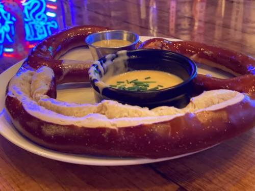 A large pretzel served with cheese dip and mustard on a plate, with a colorful neon background.
