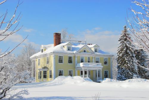 A large yellow house covered in snow, surrounded by trees and a clear blue sky.