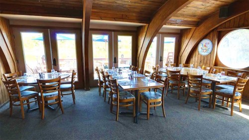 Cozy restaurant interior with wooden beams, tables set for dining, and large windows overlooking an outdoor patio.