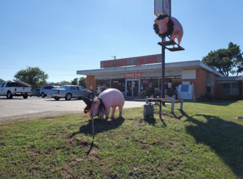 A roadside store with a sign featuring a pig, surrounded by grass and parked cars under a clear blue sky.