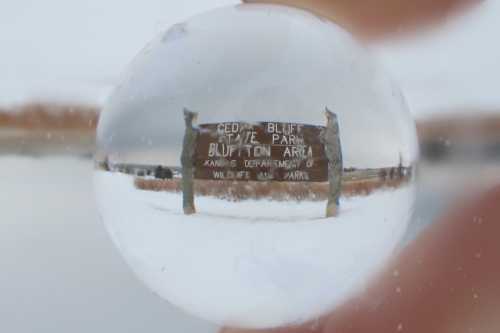 A snow-covered sign for Cedar Bluff State Park, viewed through a glass orb, creating a distorted reflection.