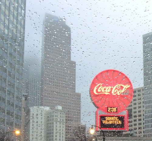 A foggy cityscape with a Coca-Cola sign displaying "DONATE, VOLUNTEER" and raindrops on the glass in the foreground.