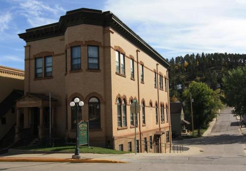 Historic brick building with large windows, located on a sloped street, surrounded by trees and a clear blue sky.