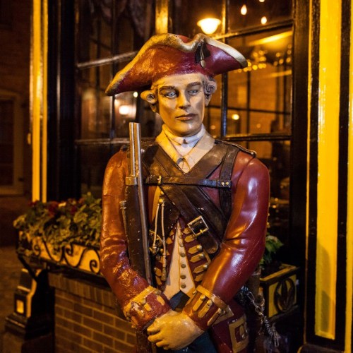 A life-sized statue of a colonial soldier in a tricorn hat, standing outside a building with warm lighting.