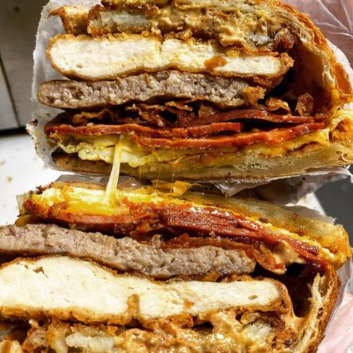 Close-up of a layered sandwich with crispy chicken, beef, cheese, and sauces between toasted bread slices.
