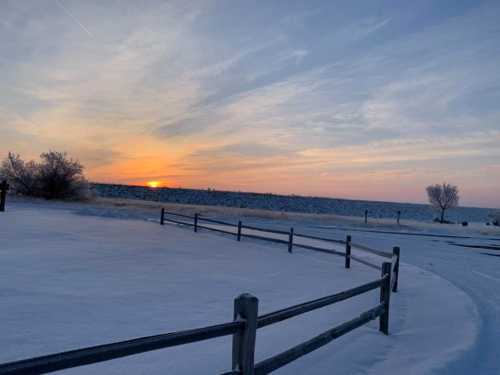 A serene winter landscape at sunset, with snow-covered ground and a wooden fence against a colorful sky.