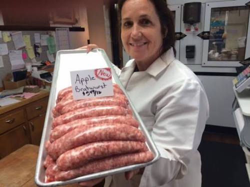 A woman in a white coat holds a tray of apple bratwurst with a sign displaying the price.