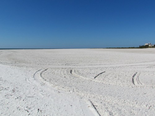 A wide, sandy beach with white sand and clear blue skies, featuring tire tracks leading towards the water.