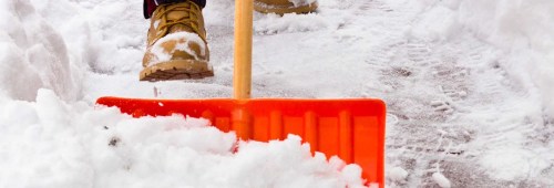 A close-up of a boot stepping on snow while using an orange snow shovel to clear a path.