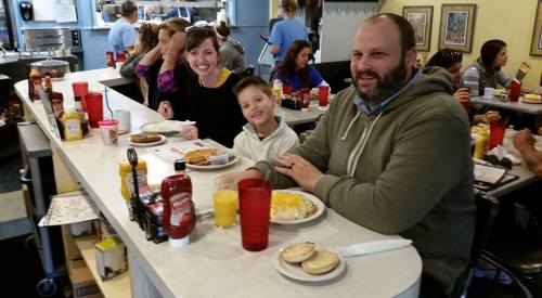 A family enjoys breakfast at a diner, smiling and sitting at a counter with food and drinks in front of them.