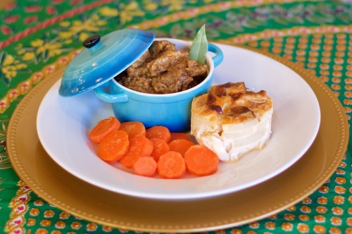 A blue pot with beef stew, served with sliced carrots and a creamy pastry on a decorative plate.