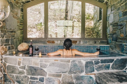 A person relaxes in a stone bathtub, enjoying wine with candles, surrounded by nature through large windows.