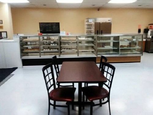 A small dining area with a table and chairs, facing a display case filled with baked goods in a bright, empty café.