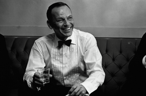 A smiling man in a tuxedo holds a glass, seated on a tufted couch, exuding a relaxed and joyful demeanor.