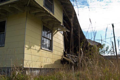 An abandoned yellow house with a sagging roof, surrounded by tall grass and under a blue sky.