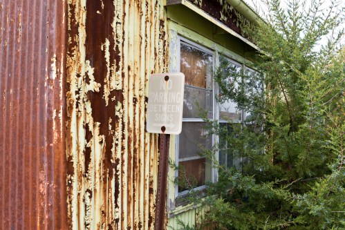A weathered building with peeling paint and a "No Parking Between Signs" sign partially obscured by greenery.
