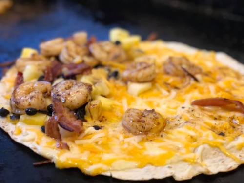 A close-up of a tortilla topped with shrimp, cheese, pineapple, and black beans, sizzling on a grill.
