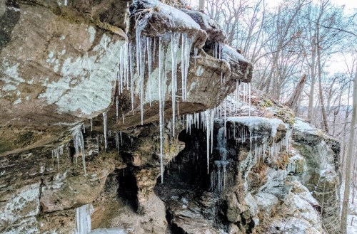 Icicles hang from a rocky overhang, surrounded by bare trees in a winter landscape.