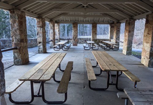 A spacious, empty picnic shelter with wooden tables and stone pillars, surrounded by trees.