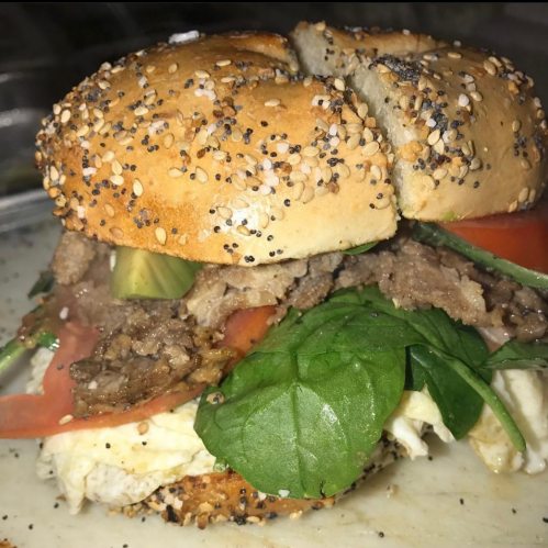 A close-up of a sesame seed bagel sandwich filled with beef, spinach, tomato, and pickles.
