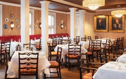 Elegant restaurant interior with wooden chairs, white tablecloths, and decorative wall plates. Warm lighting and artwork enhance ambiance.