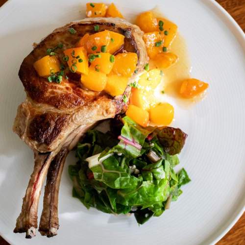 A grilled pork chop topped with diced peaches, served with a side of mixed greens salad on a white plate.