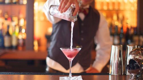 A bartender pours a pink cocktail into a martini glass at a bar, with bottles in the background.