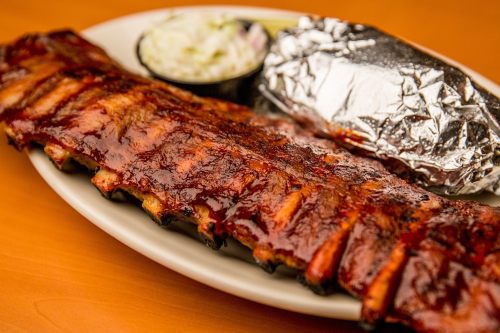 A plate of barbecue ribs glazed with sauce, served with coleslaw and a foil-wrapped baked potato.