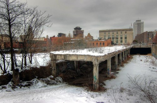 Abandoned concrete structure with snow, surrounded by overgrown vegetation and urban buildings under a cloudy sky.