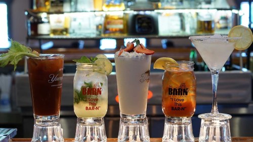 Four colorful cocktails displayed on a bar, featuring garnishes like lemon and strawberries in branded glasses.