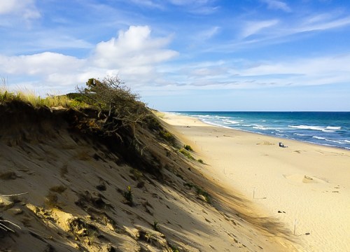 A sandy beach with gentle waves, dunes, and a blue sky with scattered clouds.