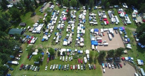 Aerial view of a large outdoor event with numerous tents, vehicles, and people in a green field surrounded by trees.