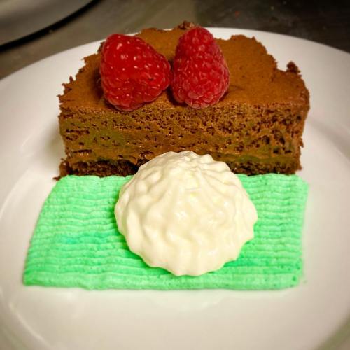 A slice of chocolate mousse cake topped with two raspberries, served with a dollop of whipped cream on a green plate.