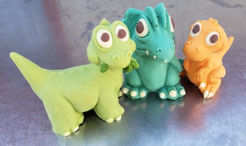 Three colorful dinosaur figurines: green, teal, and orange, with big eyes and playful expressions, made from modeling clay.